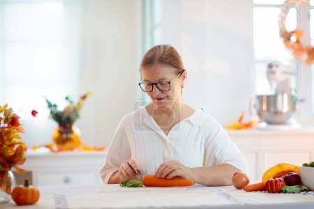 Photo for Thanksgiving dinner preparation. Woman cutting pumpkin, carrot and onion for autumn season pie. Decorated kitchen in Halloween season. Female cooking celebration feast. Seasonal vegetables. - Royalty Free Image
