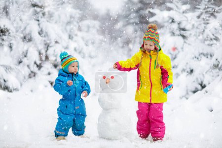 Foto de Los niños hacen muñeco de nieve. Niños construyendo muñeco de nieve jugando al aire libre en el soleado día de invierno nevado. Diversión familiar al aire libre en vacaciones de Navidad. Niño y niña juegan bolas de nieve. Ropa de invierno para bebés y niños pequeños
. - Imagen libre de derechos
