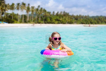 Téléchargez les photos : Enfant avec anneau gonflable sur la belle plage. Petite fille nageant dans la mer exotique. Vacances en mer avec un enfant. Les enfants jouent sur la plage d'été. Amusant. Les enfants nagent. Vacances en famille sur l'île tropicale. - en image libre de droit