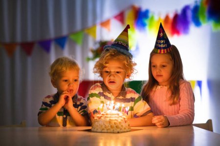 Photo for Kids birthday party. Children blow out candles on cake in dark room. Rainbow decoration and table setting for kids event, banner and flag. Girl and boy with birthday presents. Family celebration. - Royalty Free Image