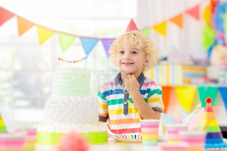 Téléchargez les photos : Fête d'anniversaire des enfants. Enfant soufflant des bougies sur un gâteau coloré. Maison décorée avec bannières drapeau arc-en-ciel, ballons. Fête des animaux de ferme thème. Petit garçon fêtant son anniversaire. Nourriture de fête
. - en image libre de droit