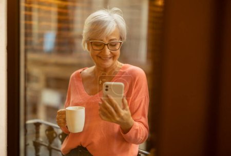 Photo for Happy senior Caucasian woman in apartment on balcony, drinking coffee while checking her cell phone - Royalty Free Image