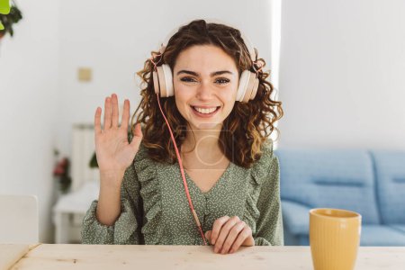 Téléchargez les photos : Portrait de souriante jeune femme caucasienne avec écouteurs agitant la main en regardant la caméra - en image libre de droit