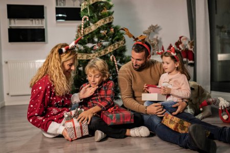 Happy caucasian family of four opening Christmas presents on the floor near decorated tree