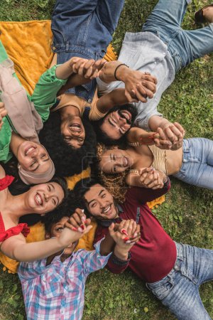 Foto de Grupo multirracial de jóvenes con los brazos levantados y las manos unidas tumbadas en la hierba - Imagen libre de derechos