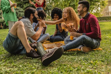 Multiracial group of friends sitting on the grass on park eating pizza and drinking juice. Focus on person with leg prosthesis and African woman.