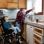 Hispanic mother looking through the window in the kitchen with her disabled son in a wheelchair. Family, unconditional love and inclusion concept.
