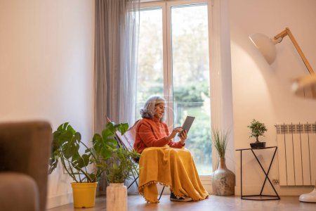 Téléchargez les photos : Old gray-haired woman resting at home with a tablet in her hand, by the window. take a blanket because it is cold. home life. - en image libre de droit
