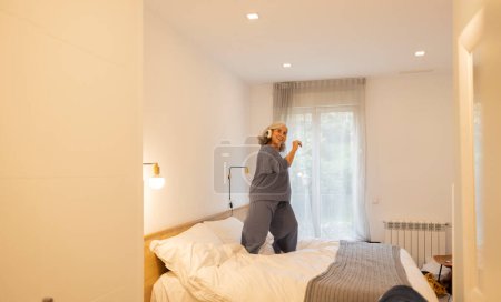 Téléchargez les photos : Exuberant mature woman jumping on the bed - listening to music and dancing at home happily - - en image libre de droit