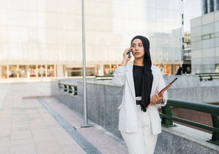 Téléchargez les photos : Femme d'affaires musulmane, dans la ville, avec un dossier et un téléphone portable parlant affaires. - en image libre de droit