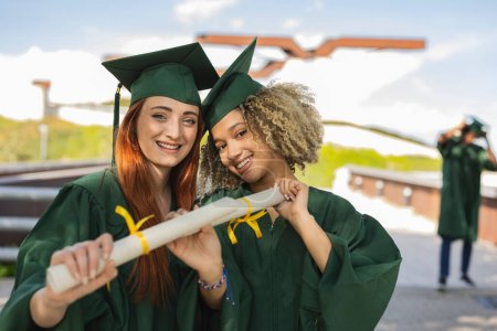 Photo for Two multiracial college girl friends at graduation with diplomas, on campus - Royalty Free Image