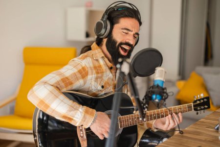 Photo for Performing song. Inspired man, amateur music singer and writer, sitting in headphones, with guitar in at home sound recording studio. Young guitarist sings and plays live instrumental melody - Royalty Free Image