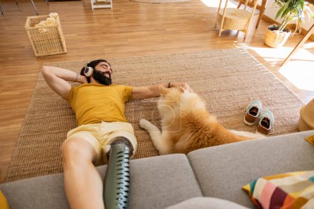 Téléchargez les photos : Homme avec prothèse de jambe amputée, à la maison avec son chien, assis sur le tapis et le canapé tout en écoutant de la musique - en image libre de droit
