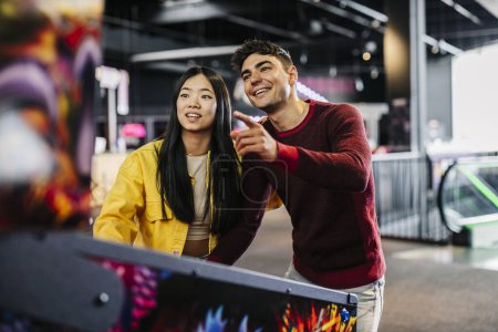 Photo for Generation z multiracial couple playing pinball machine in the game room - Royalty Free Image