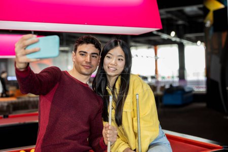 Young couple taking a selfie with pool cues at an indoor game zone.