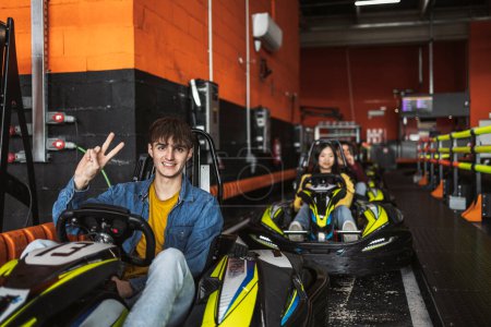 Smiling young man gives a peace sign before starting a go-kart race, with a focused female driver in the background.
