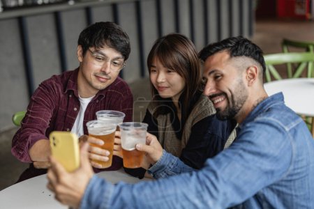 Three friends toast with beers while taking a selfie at an urban bar.