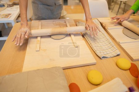 Detaillierte Ansicht von geschickten Händen, die Ton ausrollen, wobei in einer Werkstatt Töpferwerkzeuge bereit stehen.