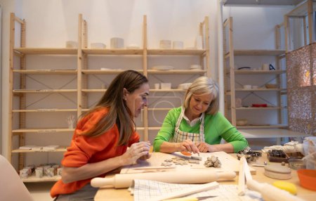 An adult and a senior woman shape clay figures in a workshop, sharing a moment of artistic joy.