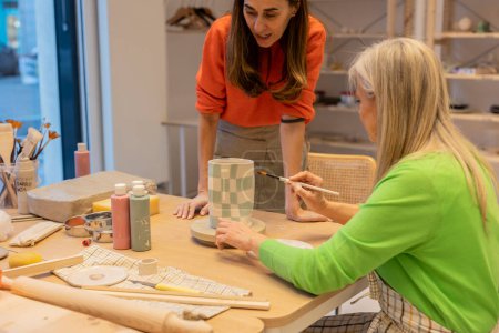 Eine erwachsene und ältere Frau malt in einer Werkstatt ein kariertes Muster auf eine Keramikvase.