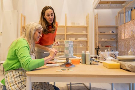 A focused session of two women painting geometric patterns on a ceramic vase at a craft workshop.