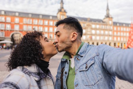 Un moment intime comme un jeune couple partage un baiser, capturant un selfie dans une place de la ville.