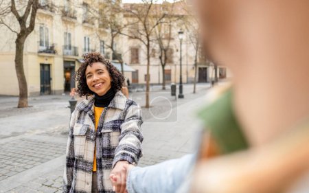Cheerful woman leads her partner by hand through a quaint city street, smiling back.