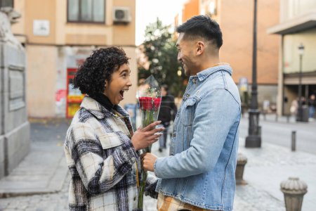 Excited young woman receives a surprise bouquet of red roses from her partner on a city sidewalk.
