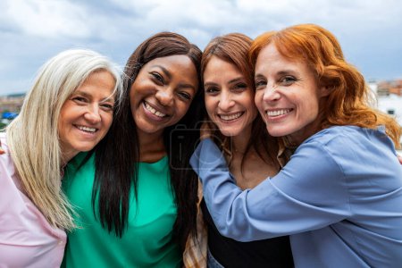 Cuatro mujeres vibrantes acurrucadas en un abrazo grupal, sonriendo brillantemente, con el cielo abierto detrás de ellas.