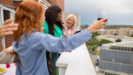 Eine fröhliche Gruppe von Frauen genießt ein Glas Wein auf einer Dachterrasse und umarmt den Blick auf die Stadt.