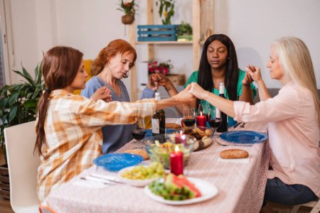 A group of women holding hands in a moment of reflection at a dinner table, surrounded by a festive setting.