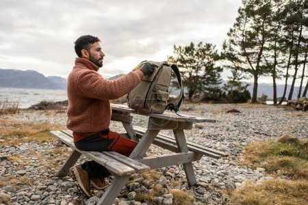 A man in a cozy sweater packs his bag at a picnic table, with a calm lake and mountains in the distance.