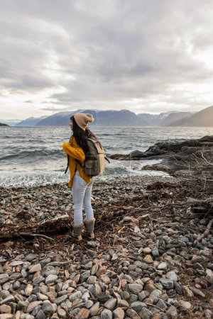 A young explorer in a knitted hat stands on a rocky beach, gazing at the expansive sea ahead.