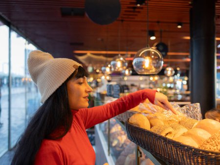 A woman carefully selects bread at an indoor market, illuminated by warm lighting.