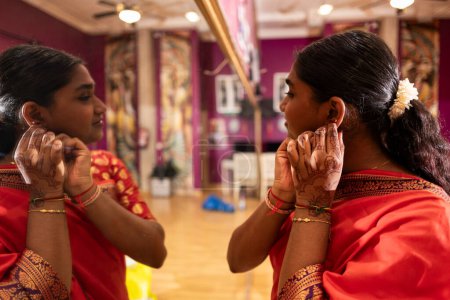 A young woman in a red saree adjusts her earrings before an event.