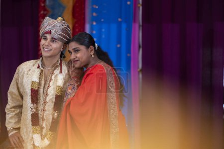 Una joven en un saree rojo se apoya en un hombre en un sherwani dorado, compartiendo un momento cultural.