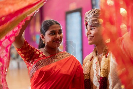 Una mujer en un sari y un hombre en un sherwani ríen juntos, rodeados de vibrantes cortinas.
