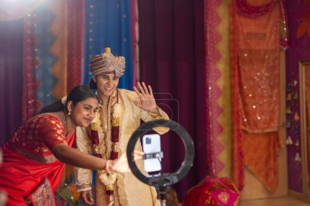 Sonriendo ampliamente, una mujer joven en un saree y un hombre en un sherwani capturan un selfie durante una festividad cultural.