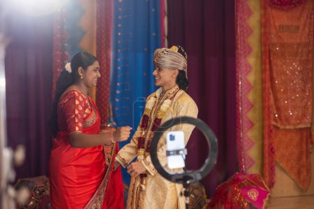 Una joven en un saree y un hombre en un sherwani intercambian saludos en una sala festivamente decorada.