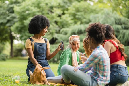 In einer üppigen Parklandschaft nutzt eine Frau ihr Smartphone, um die Freude eines Picknicks mit Freunden einzufangen.