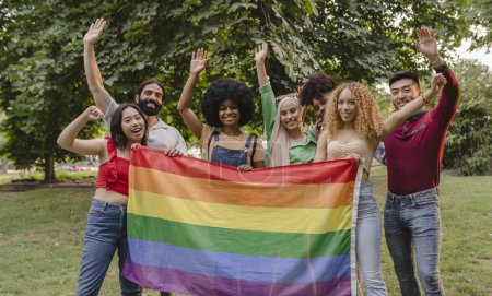 Un grupo diverso y alegre de amigos levanta sus manos en unidad, orgullosamente sosteniendo la bandera del arco iris en un parque.