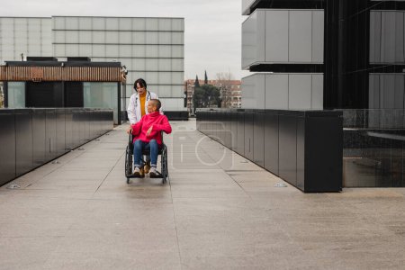 Una suave interacción entre un trabajador sanitario y una joven en silla de ruedas, compartiendo un momento en una azotea moderna.