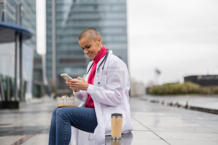 Eine glückliche Ärztin genießt einen leichten Moment auf ihrem Handy während einer Stadtlandschaftspause, Kaffee an ihrer Seite.