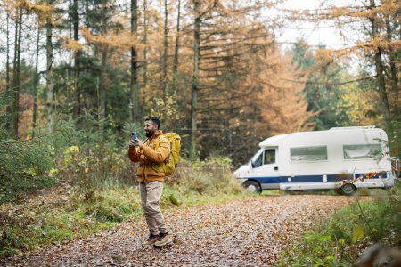 A bearded man in a yellow jacket navigates with his smartphone near a camper among autumn trees.