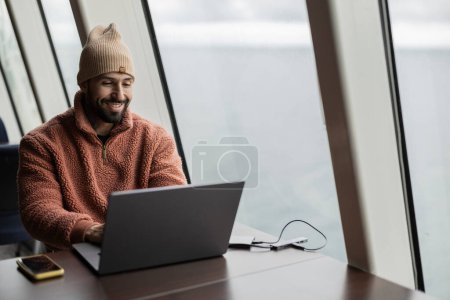 A smiling man in a beanie enjoys his work on a laptop, sitting comfortably in a modern coastal office.