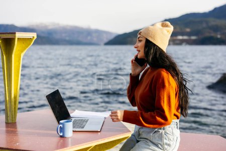 A young woman multitasking with a phone call and laptop work at a lakeside outdoor office setup.