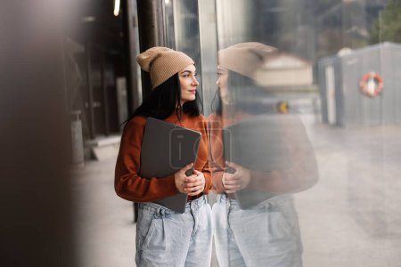 Young female entrepreneur holding a laptop, reflected on glass at an urban bus station.