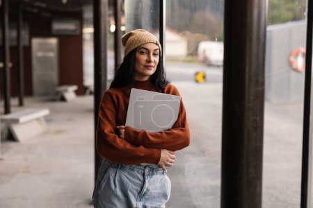 A young woman holding a laptop stands confidently at a bus station, embodying urban mobility and freelance lifestyle.