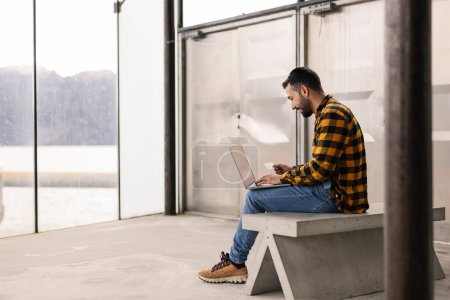 Un homme concentré dans une chemise à carreaux travaille sur son ordinateur portable dans un terminal de bus au bord du lac serein.