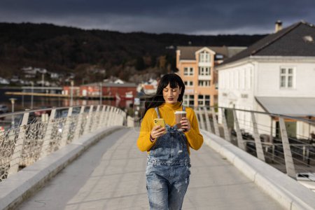 Una mujer adulta vestida de manera casual se conecta con su teléfono, paseando por un moderno puente peatonal.